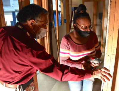 A man holds a board in place while a woman uses a drill to attach a screw to the board.