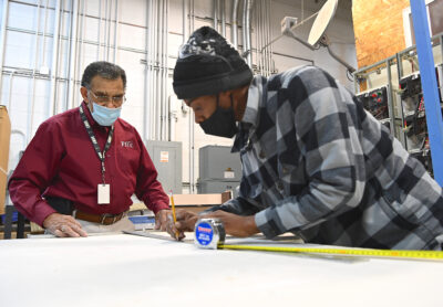 Two men lean over a piece of drywall, taking measurements.