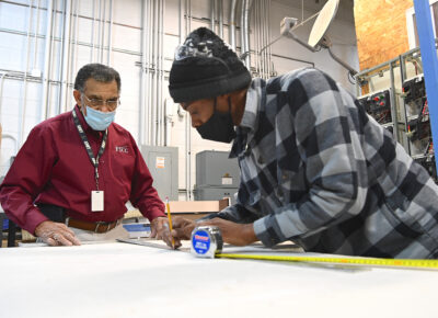 Two men lean over a piece of drywall, taking measurements.