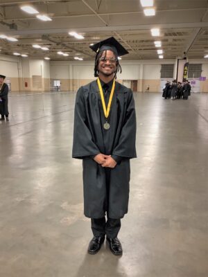 A smiling graduate stands with his feet together and his hands clasped in front of him.