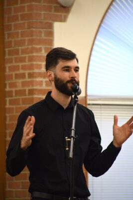 A bearded man wearing a black shirt speaks into a microphone and holds his hands out in front of him.