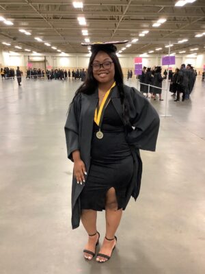 A woman with long hair and dark glasses, wearing a graduation cap, gown and a black dress stands with one hand on her hip. She is wearing a medal on a black and gold ribbon around her neck.