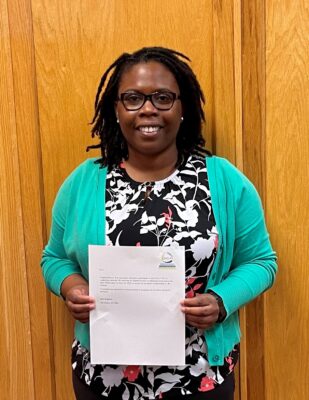 A woman wearing glasses smiles at the camera while holding a white piece of paper in her hands.