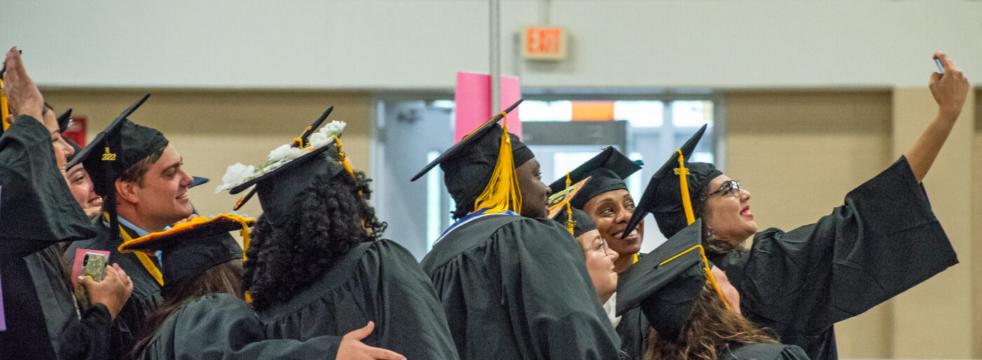 A group of graduates wearing caps and gowns pose for a selfie taken by the graduate at the front of the group.