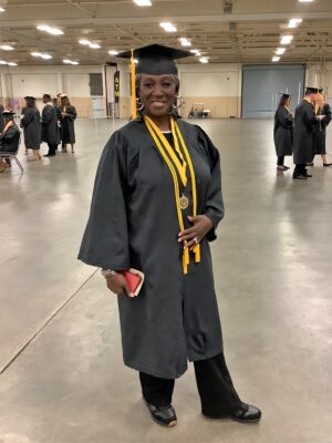 A graduate in cap and gown stands slightly turned to the side while smiling at the camera. She is wearing two gold cords and a medal on a black and gold ribbon around her neck. 