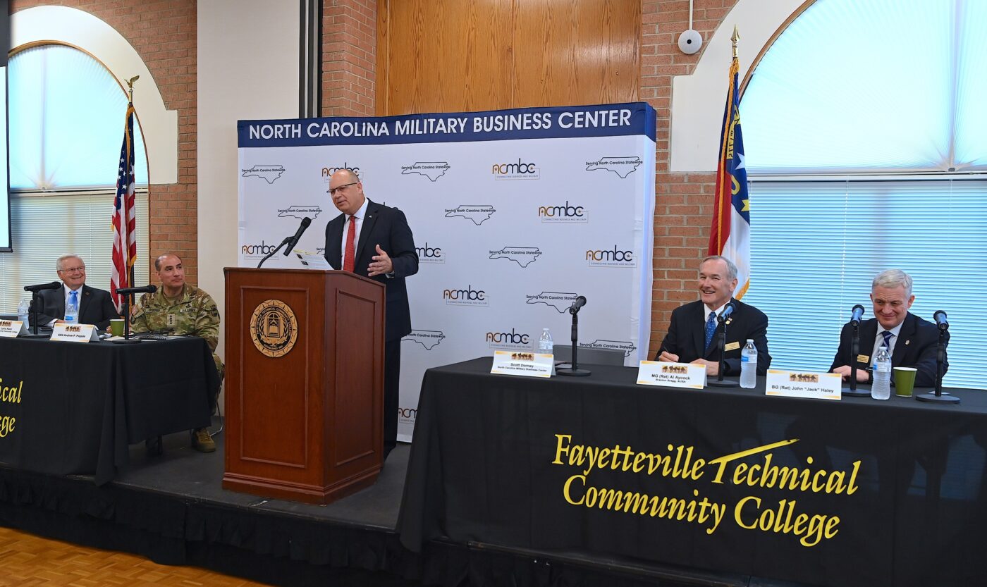 Wide photo that shows Scott Dorner speaking at the podium while other panelist sit at tables on either side of the podium onstage.