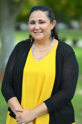 Amanda Colores, dressed in a yellow shirt and black sweater, stands outside with her hands clasped in front of her.