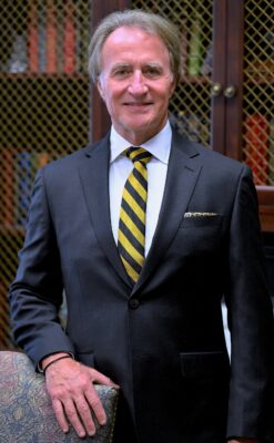 Mark Sorrells, dressed in a suit, stands with his hand on the back of a chair