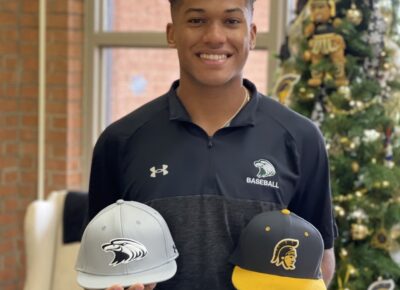 Raebert De Los Santos stands while holding a Central Methodist baseball hat in one hand and a FTCC baseball hat in the other hand.