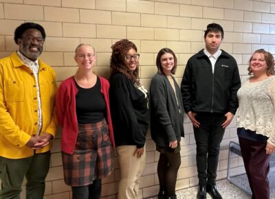 A group of five people, smiling at the camera, stand against a tile wall.