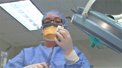 A surgical tech student dressed in operating room scrubs and protective gear cleans a pair of forceps.