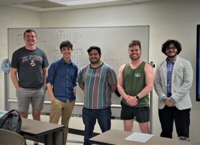 A group of males students stand in a line at the front of a classroom. A whiteboard with algebra notes written on it is visible behind them.