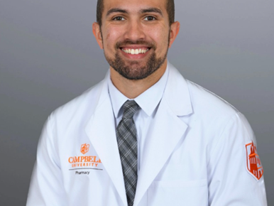 Brandon Little, with close-cropped dark hair and beard, wears a collar shirt, necktie and white pharmacist's coat.