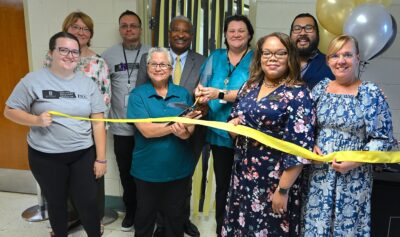 A group of people stand behind a black and yellow ribbon. One woman holds a pair of large scissors prepared to cut the ribbon.