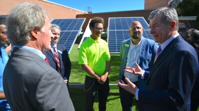 People stand around large solar panels in front of a brick building.