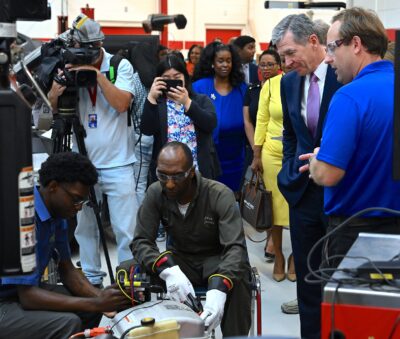 A group of people stand around watching two men work on a car part inside of an automotive shop garage.