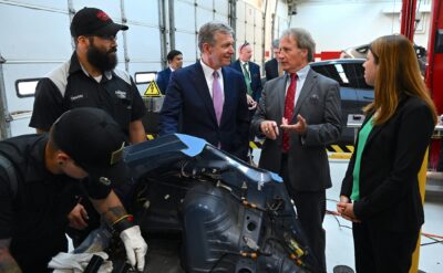 Four men, two of them in suits, and one woman stand around a piece of a car in an automotive shop garage.