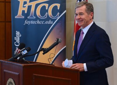 Governor Roy Cooper, dressed in a suit, stands at a podium. A FTCC banner is visible behind him.