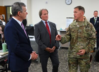 Two men in suits and one man in a military uniform stand talking in an office room.