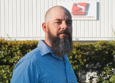 Brandon Meredith, a bald man with a long beard wearing a blue shirt, stands outside.