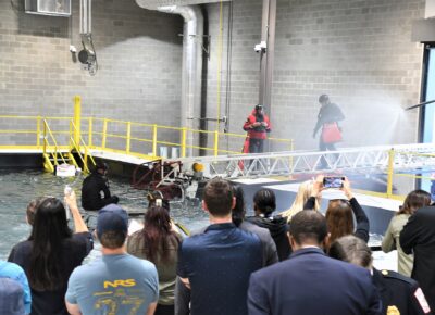 A man walks on a fire truck ladder extended over a water tank where another man sits on top of a submerged car.