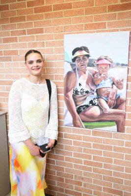 A woman stands next to a painting of a grandmother and granddaughter sitting together on a beach.