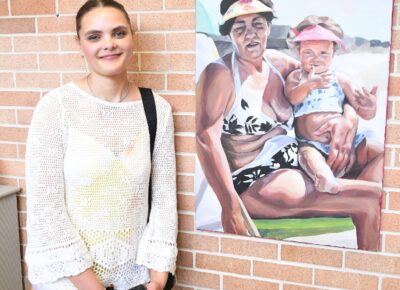 A woman stands next to a painting of a grandmother and granddaughter sitting together on a beach.