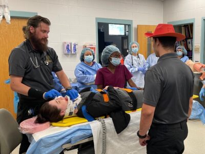 Allied Health students stand around a student pretending to be injured on an operating table in a classroom.