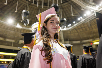 A young woman in a pink graduation cap and gown looks at the camera.