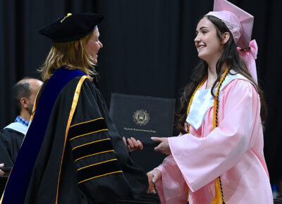 A young woman in a pink graduation cap and gown shakes hands with a woman in doctoral robes while accepting a degree.