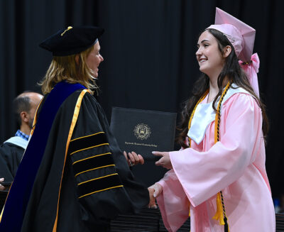 A young woman in a pink graduation cap and gown shakes hands with a woman in doctoral robes while accepting a degree.