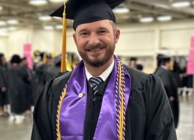 Roger Edwards stands wearing a cap and gown with a purple stole and a gold cord.