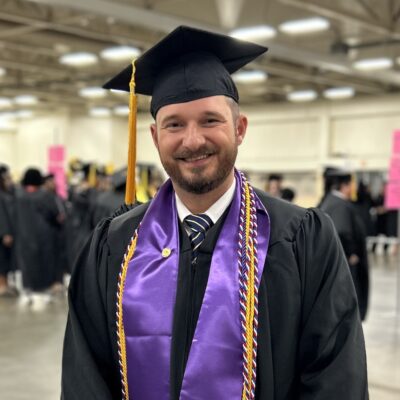 Roger Edwards stands wearing a cap and gown with a purple stole and a gold cord.