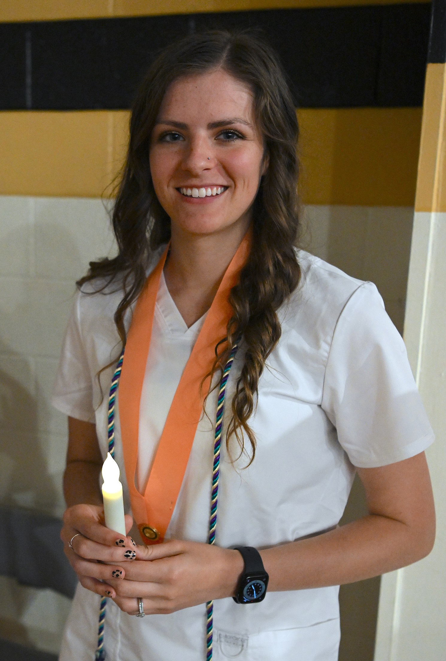 A close-up of a Nursing graduate, wearing a peach-colored pin ribbon and a cord. She is holding a battery-operated candle.