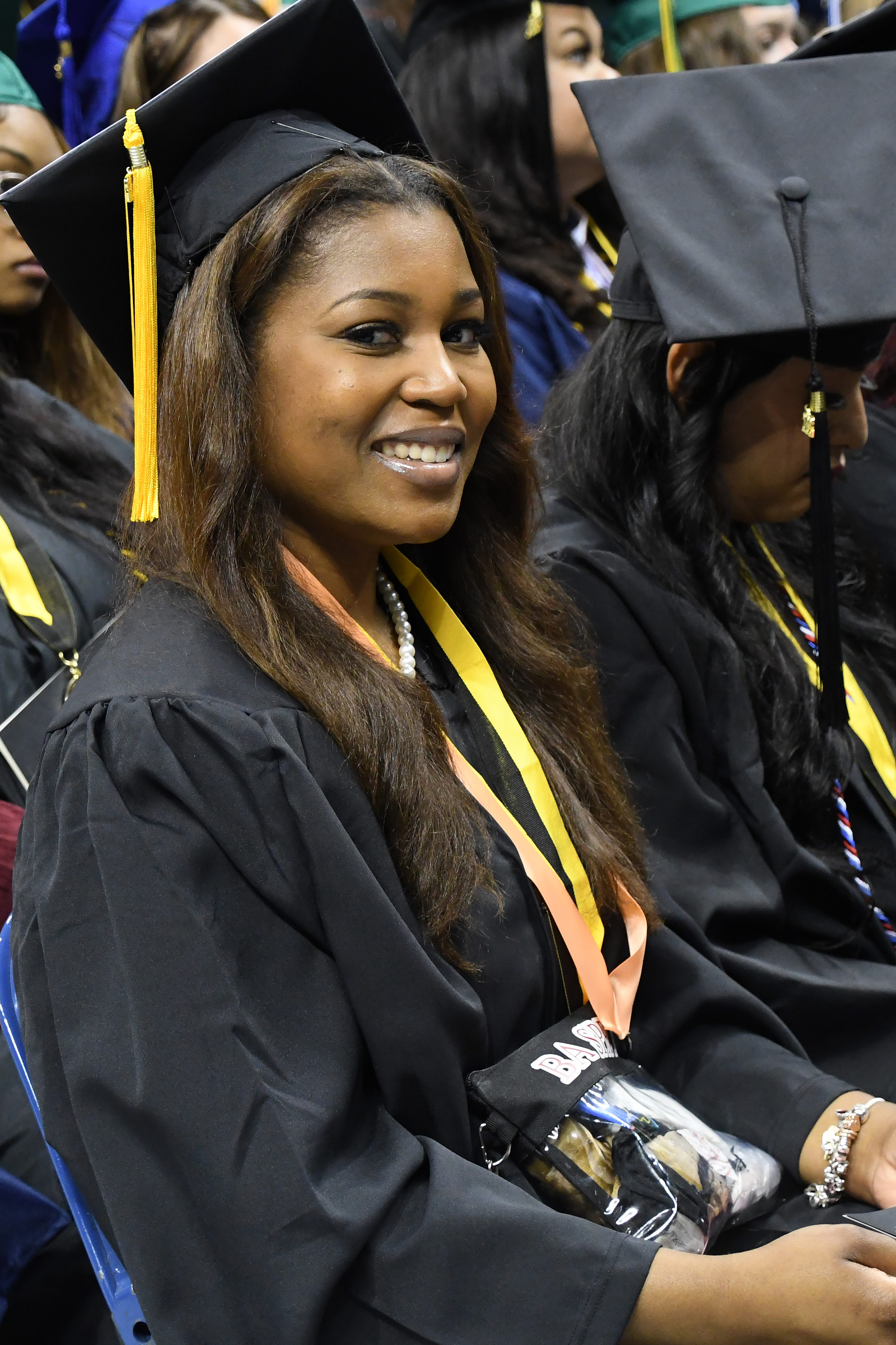 A graduate wearing a string of pearls and two ribbons around her neck smiles at the camera.