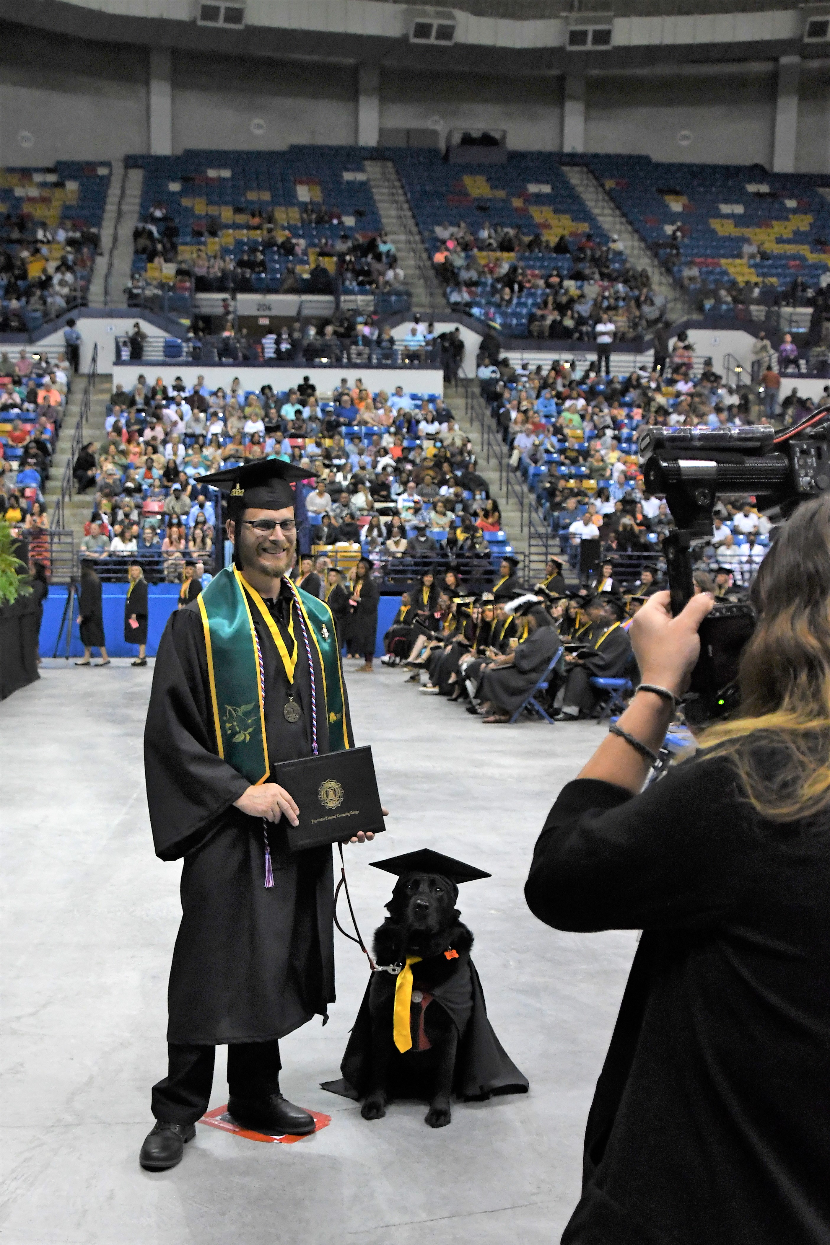 A graduate wearing a green stole and red, white and blue cord poses with his service dog, a black Labrador retriever who is also wearing a cap and gown. The human graduate is holding his degree portfolio. 