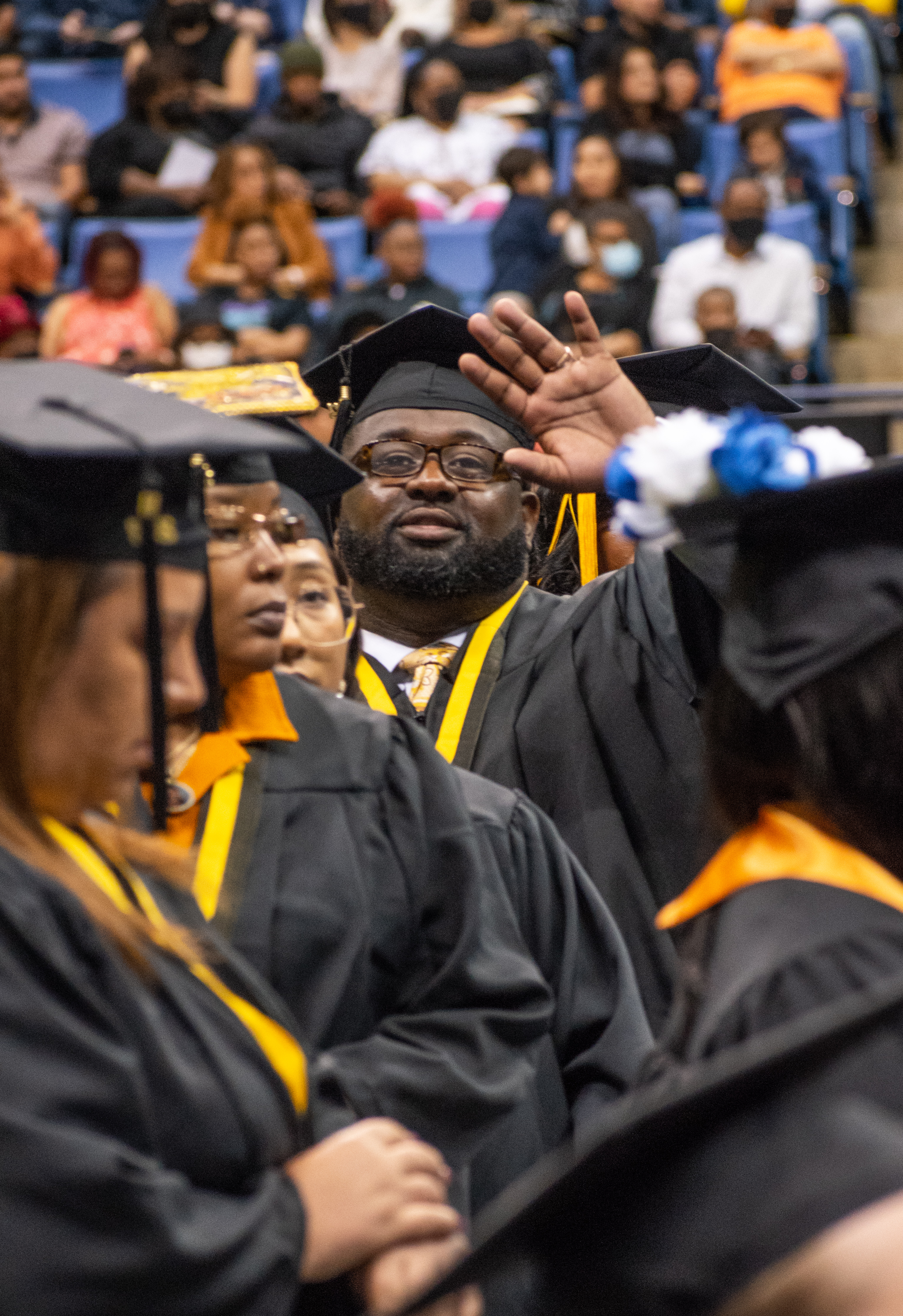 A graduate waves to the camera. He is surrounded by a crowd of graduates.