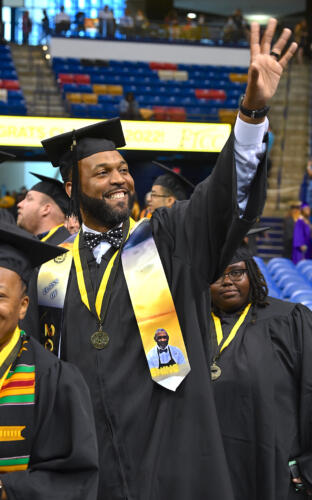A graduate, wearing a personalized gold and blue stole with the image of a man on it, smiles and waves to the crowd.