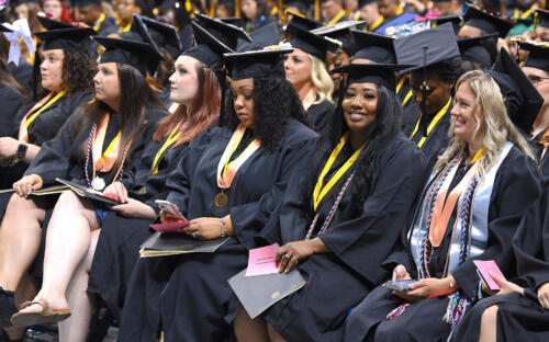 Six graduates sit in a row. The second graduate from the right is smiling at the camera.