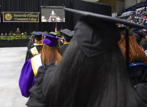 The back of several graduates as they face the stage. A larger projector screen and Fayetteville Technical Community College banner are visible in the background.