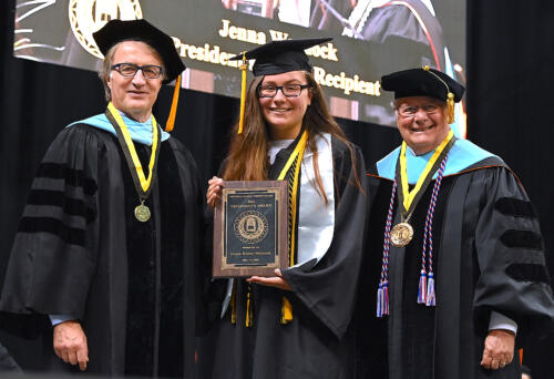 Jenna Warnock holds a plaque. She is dressed in her cap and gown. Dr. Mark Sorrells and Dr. Larry Keen stand on either side of her.