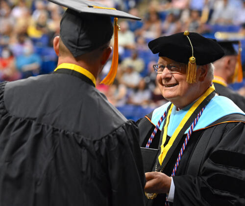 Dr. Larry Keen smiles at a graduate as he hands him his degree portfolio. The back of the graduate is in the foreground.