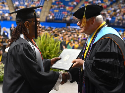 A graduate wearing glasses, earrings and a silver stole accepts a degree portfolio from Dr. Larry Keen while shaking his hand. The audience is visible in the background of the photo.