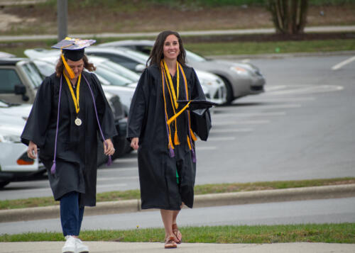 Two graduates walk up the sidewalk. One is wearing her cap, and the other is carrying her cap.