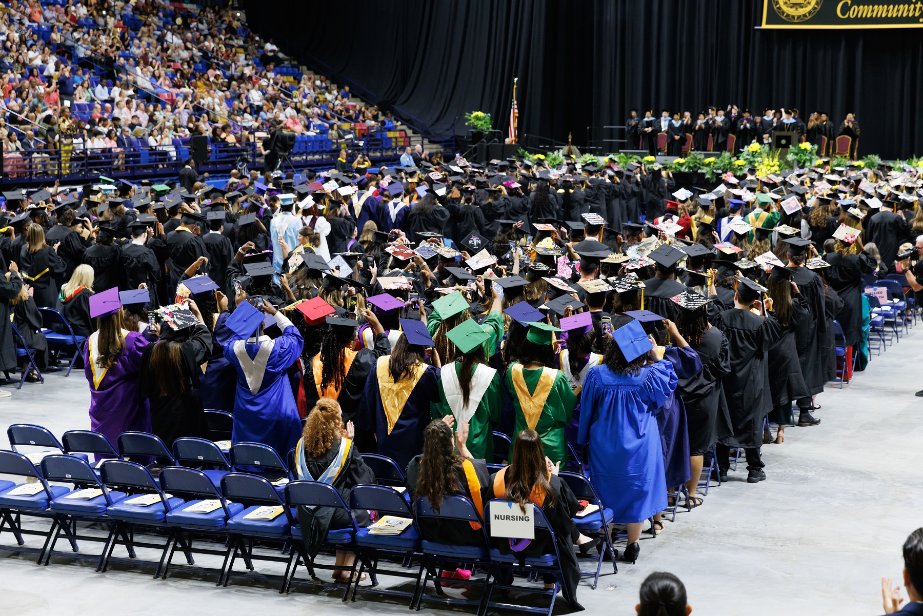 A photo taken from behind the rows of graduates. They are standing up and reaching to turn their tassels.