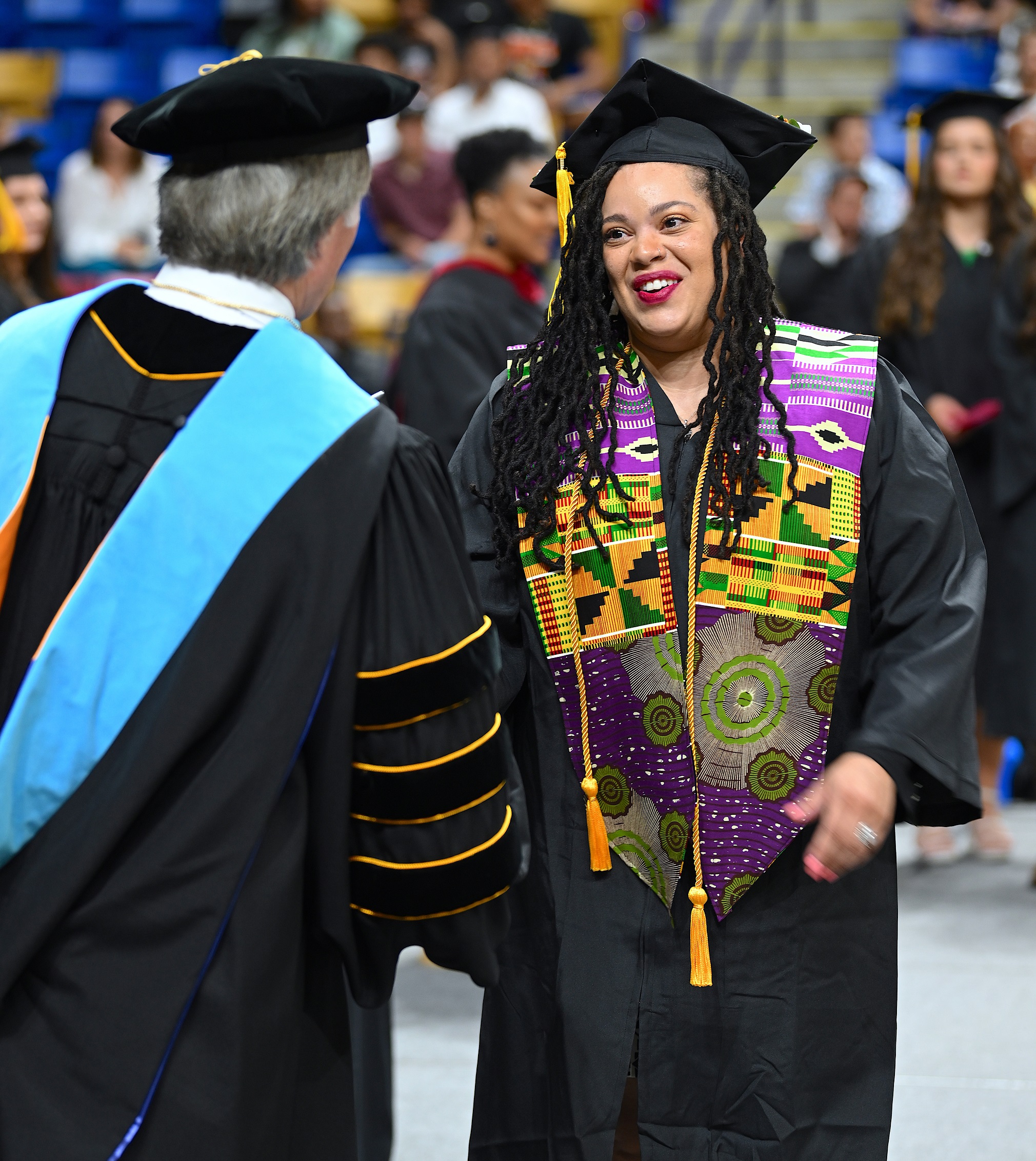 A graduate wearing a yellow, green and purple stole smiles as she shakes hands with Dr. Mark Sorrells.