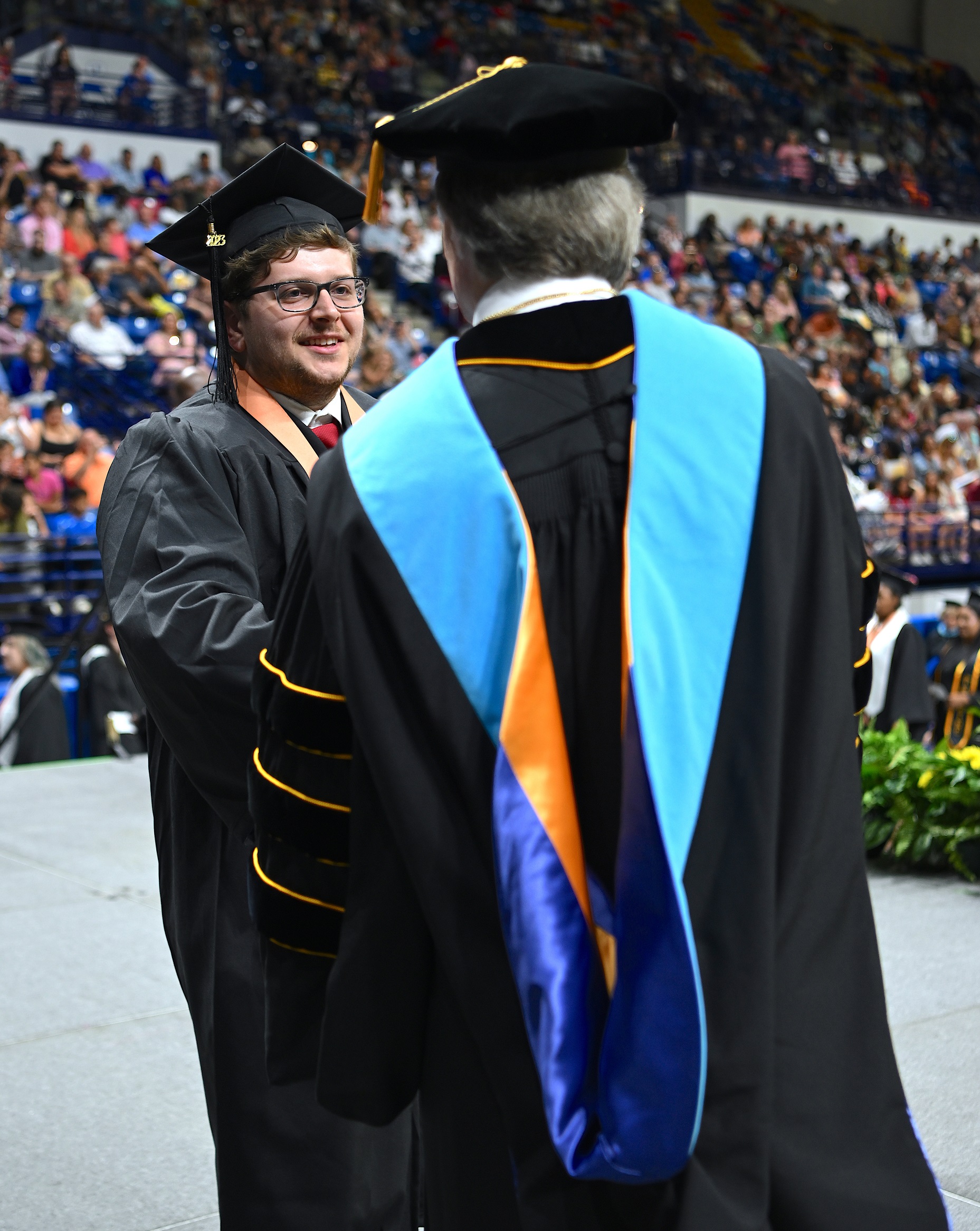 A graduate wearing a peach-colored ribbon shakes hands with Dr. Mark Sorrells, who is shown from behind.