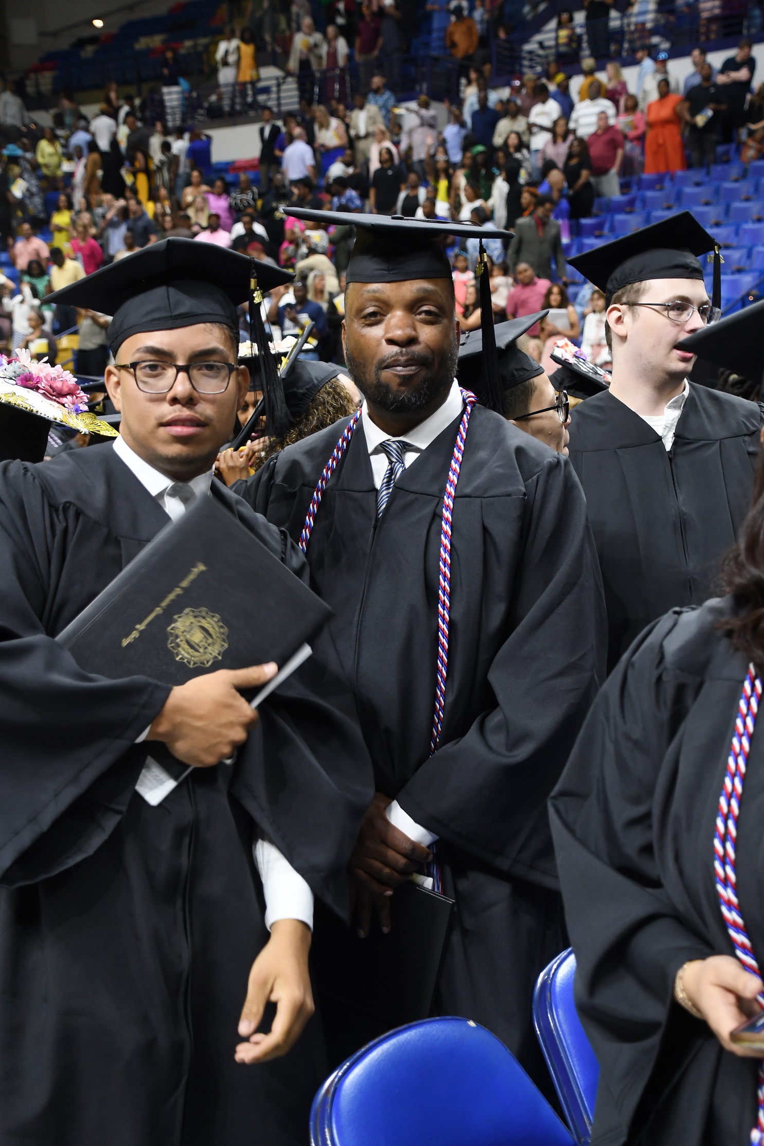 Two graduates with serious faces look at the camera. One graduate is holding his diploma folder and the other graduate is wearing a red, white and blue cord.
