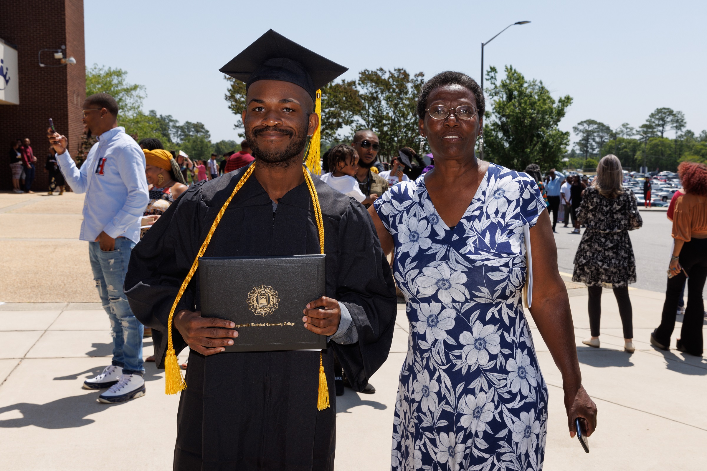 A graduate wearing his cap, gown and gold cord hold his diploma folder as he stands next to a woman.