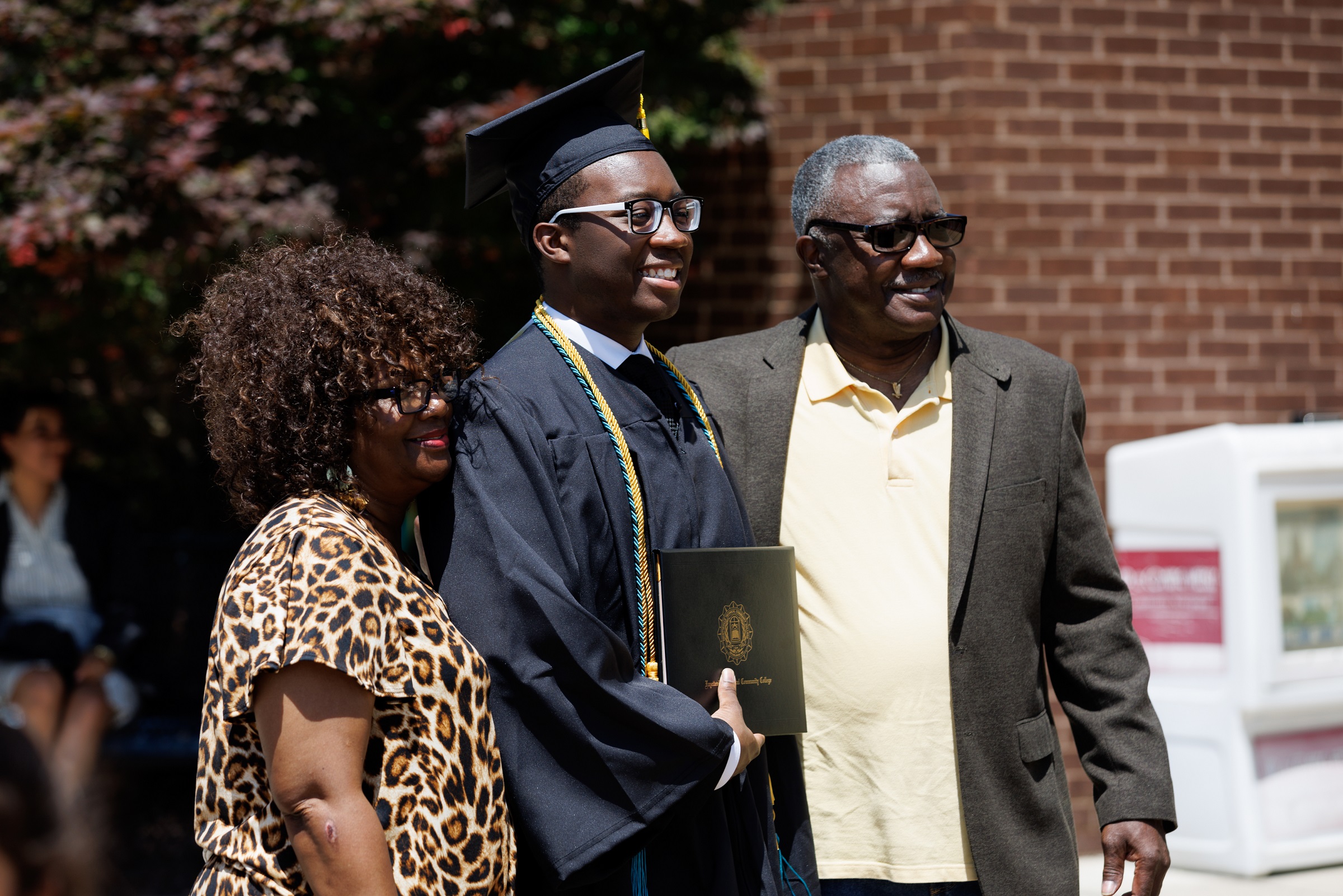 A graduate wearing glasses and two cords holds his diploma folder while standing between a man and woman outside the Crown.